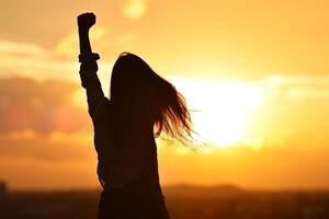 ai generado silueta de un mujer con fluido pelo con su puño elevado arriba en el puesta de sol cielo, niña poder foto