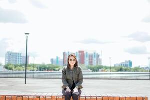 joven hermosa asiático mujer vistiendo chaqueta y negro pantalones posando al aire libre sentado en el parque foto