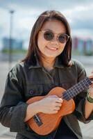 Playing Ukulele of Young Beautiful Asian Woman Wearing Jacket And Black Jeans Posing Outdoors photo