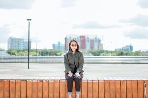 joven hermosa asiático mujer vistiendo chaqueta y negro pantalones posando al aire libre sentado en el parque foto