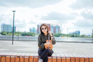 jugando ukelele de joven hermosa asiático mujer vistiendo chaqueta y negro pantalones posando al aire libre foto