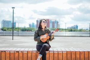 Playing Ukulele of Young Beautiful Asian Woman Wearing Jacket And Black Jeans Posing Outdoors photo