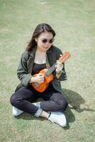 Playing Ukulele of Young Beautiful Asian Woman Wearing Jacket And Black Jeans Posing Outdoors photo