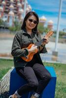 jugando ukelele de joven hermosa asiático mujer vistiendo chaqueta y negro pantalones posando al aire libre foto