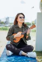 Playing Ukulele of Young Beautiful Asian Woman Wearing Jacket And Black Jeans Posing Outdoors photo