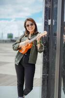 jugando ukelele de joven hermosa asiático mujer vistiendo chaqueta y negro pantalones posando al aire libre foto