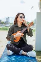 jugando ukelele de joven hermosa asiático mujer vistiendo chaqueta y negro pantalones posando al aire libre foto