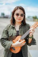 Playing Ukulele of Young Beautiful Asian Woman Wearing Jacket And Black Jeans Posing Outdoors photo