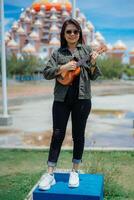 jugando ukelele de joven hermosa asiático mujer vistiendo chaqueta y negro pantalones posando al aire libre foto