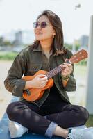 Playing Ukulele of Young Beautiful Asian Woman Wearing Jacket And Black Jeans Posing Outdoors photo