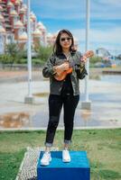 jugando ukelele de joven hermosa asiático mujer vistiendo chaqueta y negro pantalones posando al aire libre foto