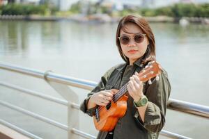 jugando ukelele de joven hermosa asiático mujer vistiendo chaqueta y negro pantalones posando al aire libre foto