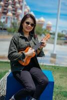 Playing Ukulele of Young Beautiful Asian Woman Wearing Jacket And Black Jeans Posing Outdoors photo