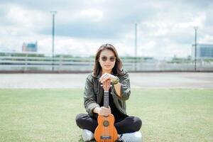 Playing Ukulele of Young Beautiful Asian Woman Wearing Jacket And Black Jeans Posing Outdoors photo