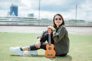 Playing Ukulele of Young Beautiful Asian Woman Wearing Jacket And Black Jeans Posing Outdoors photo