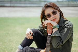 Playing Ukulele of Young Beautiful Asian Woman Wearing Jacket And Black Jeans Posing Outdoors photo