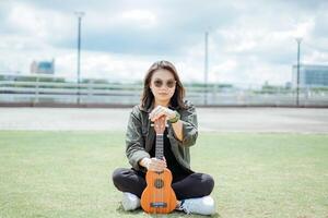 Playing Ukulele of Young Beautiful Asian Woman Wearing Jacket And Black Jeans Posing Outdoors photo