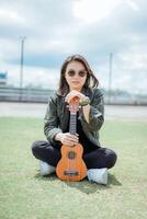 Playing Ukulele of Young Beautiful Asian Woman Wearing Jacket And Black Jeans Posing Outdoors photo