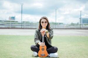 Playing Ukulele of Young Beautiful Asian Woman Wearing Jacket And Black Jeans Posing Outdoors photo