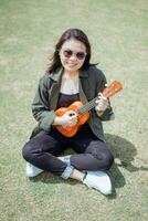 Playing Ukulele of Young Beautiful Asian Woman Wearing Jacket And Black Jeans Posing Outdoors photo