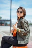Playing Ukulele of Young Beautiful Asian Woman Wearing Jacket And Black Jeans Posing Outdoors photo