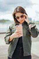 utilizando teléfono inteligente de joven hermosa asiático mujer vistiendo chaqueta y negro pantalones posando al aire libre foto