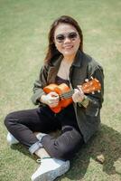 jugando ukelele de joven hermosa asiático mujer vistiendo chaqueta y negro pantalones posando al aire libre foto