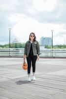 jugando ukelele de joven hermosa asiático mujer vistiendo chaqueta y negro pantalones posando al aire libre foto