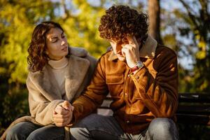 joven Pareja es sentado en parque en soleado día. hombre es triste y mujer en consolando a él. foto