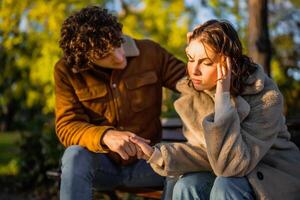 joven Pareja es sentado en parque en soleado día. mujer es triste y hombre en consolando su. foto