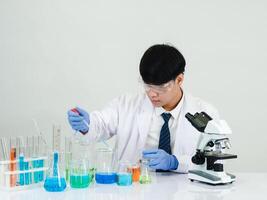 retrato asiático hombre estudiante científico vistiendo un médico vestido en el laboratorio mirando mano a químico. causado por mezcla reactivos en científico investigación laboratorios con prueba tubos y microscopio en el mesa foto