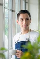 retrato jardinero adulto joven asiático hombre uno persona hermoso en pie pensar Mira y sonrisa relajarse contento mano participación Bebiendo taza café en habitación café con vaso ventana y verde árbol en Mañana tiempo. foto