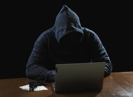 Portrait hacker spy man one person in black hoodie sitting on table looking computer laptop used login password attack security to data digital internet network system night dark background copy space photo