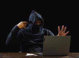 Portrait hacker spy man one person in black hoodie sitting on table looking computer laptop used login password attack security to data digital internet network system night dark background copy space photo