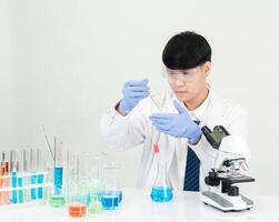 retrato asiático hombre estudiante científico vistiendo un médico vestido en el laboratorio mirando mano a químico. causado por mezcla reactivos en científico investigación laboratorios con prueba tubos y microscopio en el mesa foto