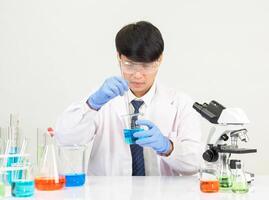 retrato asiático hombre estudiante científico vistiendo un médico vestido en el laboratorio mirando mano a químico. causado por mezcla reactivos en científico investigación laboratorios con prueba tubos y microscopio en el mesa foto