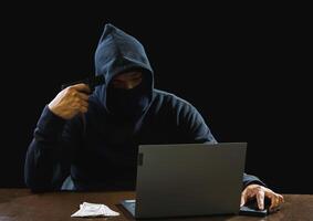 Portrait hacker spy man one person in black hoodie sitting on table looking computer laptop used login password attack security to data digital internet network system night dark background copy space photo