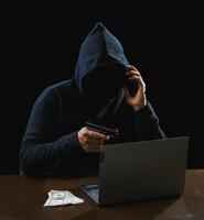 Portrait hacker spy man one person in black hoodie sitting on table looking computer laptop used login password attack security to data digital internet network system night dark background copy space photo