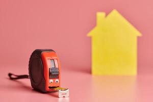 Metal tape measure funny concept. House renovation. Home repair and redecorated concept. Yellow house shaped figure on pink background. photo