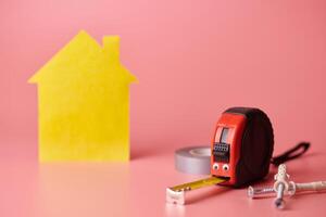 House renovation funny concept. Metal tape measure and other repair items. Home repair and redecorated concept. Yellow house shaped figure on pink background. photo