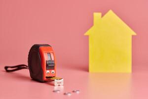 Metal tape measure funny concept. House renovation. Home repair and redecorated concept. Yellow house shaped figure on pink background. photo