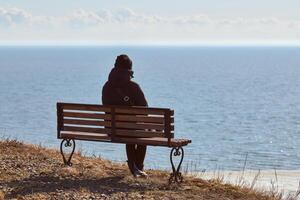 una chica soltera con una chaqueta negra y un sombrero sentada en un banco en un acantilado frente al mar, un lugar tranquilo y tranquilo foto