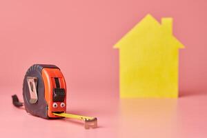 Metal tape measure funny concept. House renovation. Home repair and redecorated concept. Yellow house shaped figure on pink background. photo