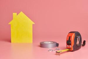 House renovation funny concept. Metal tape measure and other repair items. Home repair and redecorated concept. Yellow house shaped figure on pink background. photo