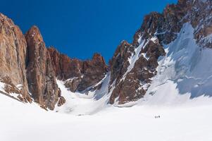 A group of climbers is visible against the backdrop of majestic rocky peaks covered with snow under a clear blue sky. The steep slopes and extreme terrain make it a challenging yet popular destination photo