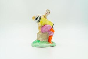 Antique ceramic musician figure, seated accordion player. Porcelain glossy finish. Traditional attire, bright orange trousers, pink waist sash, green rounded base, folk hand-painted. White background photo