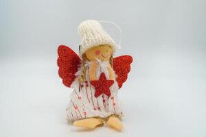 Handmade knitted Christmas angel doll, red glitter wings, holding red star, adorned with white hat, blond yarn hair, heart-printed dress, golden feet, smiling wooden face. White background photo
