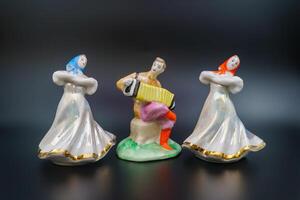 Ensemble, porcelain figurines, ceramic girls flanking accordion player. Dresses in motion, traditional attire, draped headscarves, folk hand-painted, craftsmanship. Festive assembly. Black background photo