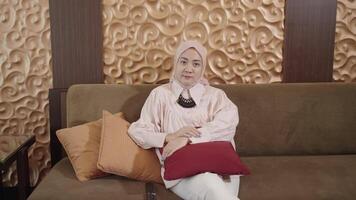 moderno musulmán mujer en hijab Sureste asiático - acecho televisión utilizando remoto mientras sentado en sofá en el vivo habitación video
