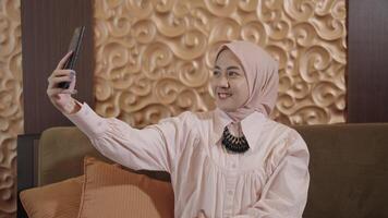 modern Muslim Frau auf Hijab Süd-Ost asiatisch - - lächelnd, Pose, nehmen Selfie Foto mit ihr Smartphone während Sitzung auf Couch im das Leben Zimmer video